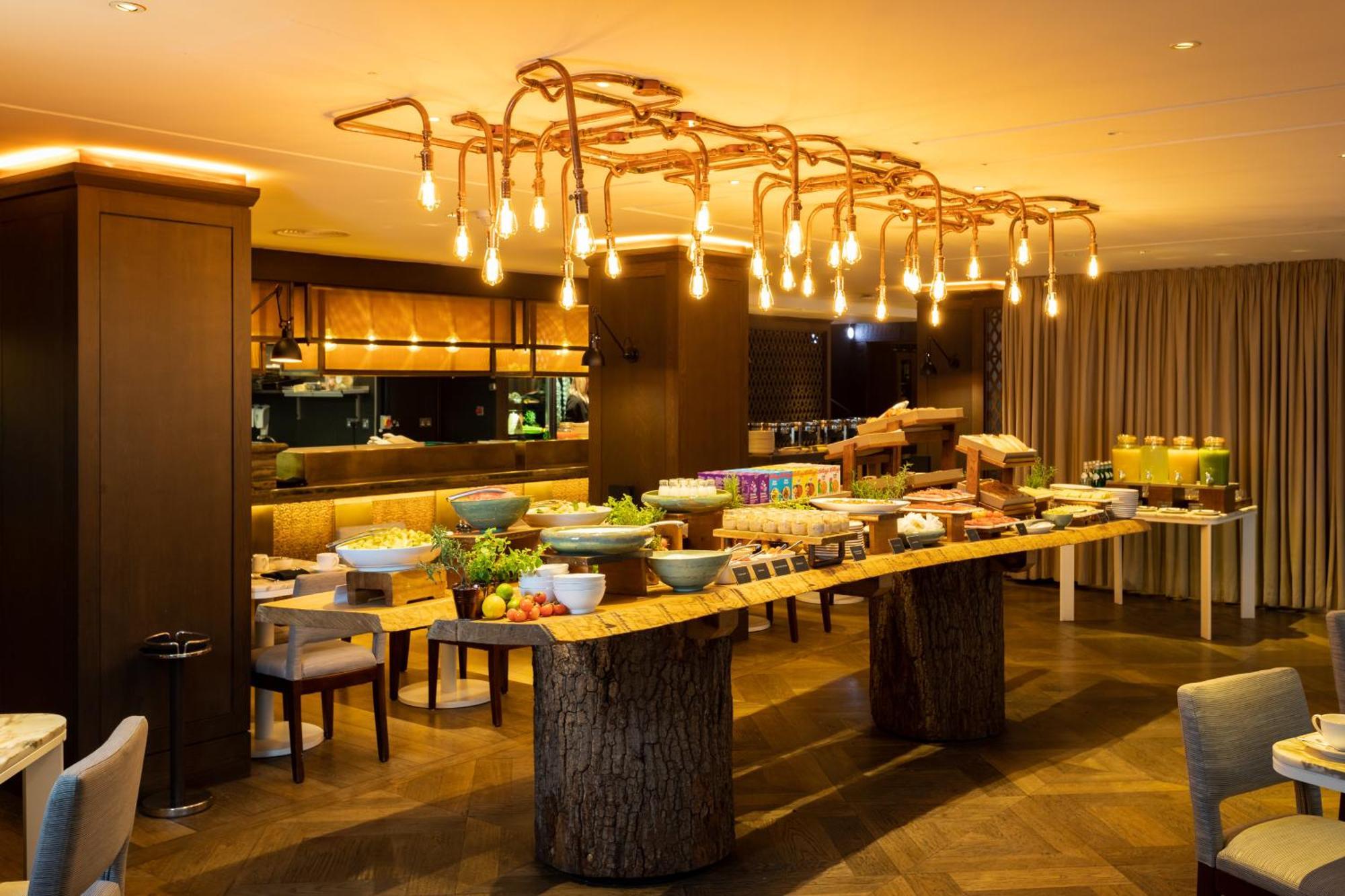 The May Fair, A Radisson Collection Hotel, Mayfair London Luaran gambar The photo shows a stylish dining area with a buffet setup. In the center, there is a long wooden table made from a tree trunk, displaying various dishes and food items. The setting features an elegant lighting fixture made of exposed bulbs that resem